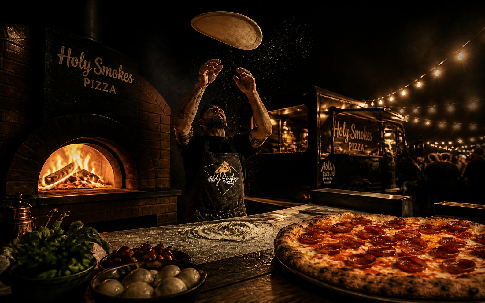 Holy Smokes Pizza open kitchen: wood-fired oven, pizza maker tossing dough, and food trailer under string lights